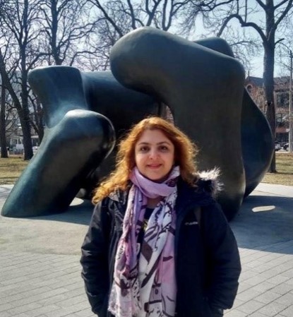 Picture of woman with brown hair wearing a scarf and standing in front of a large sculpture