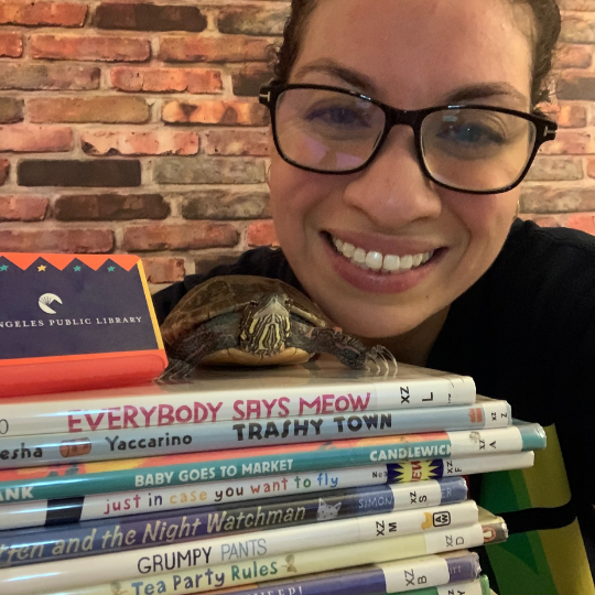 Lauren Kratz Prushko is smiling behind a large stack of children's book. A small turtle is on top of the book stack.