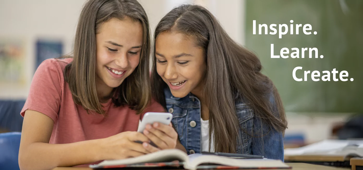 Photo of two smiling girls sitting at a desk, looking at a smartphone together. Open books are on the desk in front of them. The background features a chalkboard, and to the right, the text 'Inspire. Learn. Create.' is displayed.