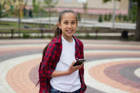 A young girl standing in the park, holding a mobile phone, looking into the camera and smiling